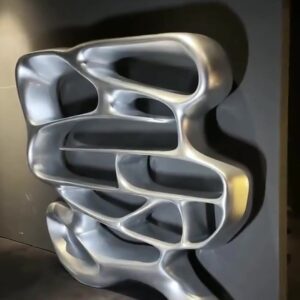 Full view of a freestanding organic-shaped mirror-polished stainless steel bookshelf with five tiers of curved openings, standing against a dark grey gallery wall