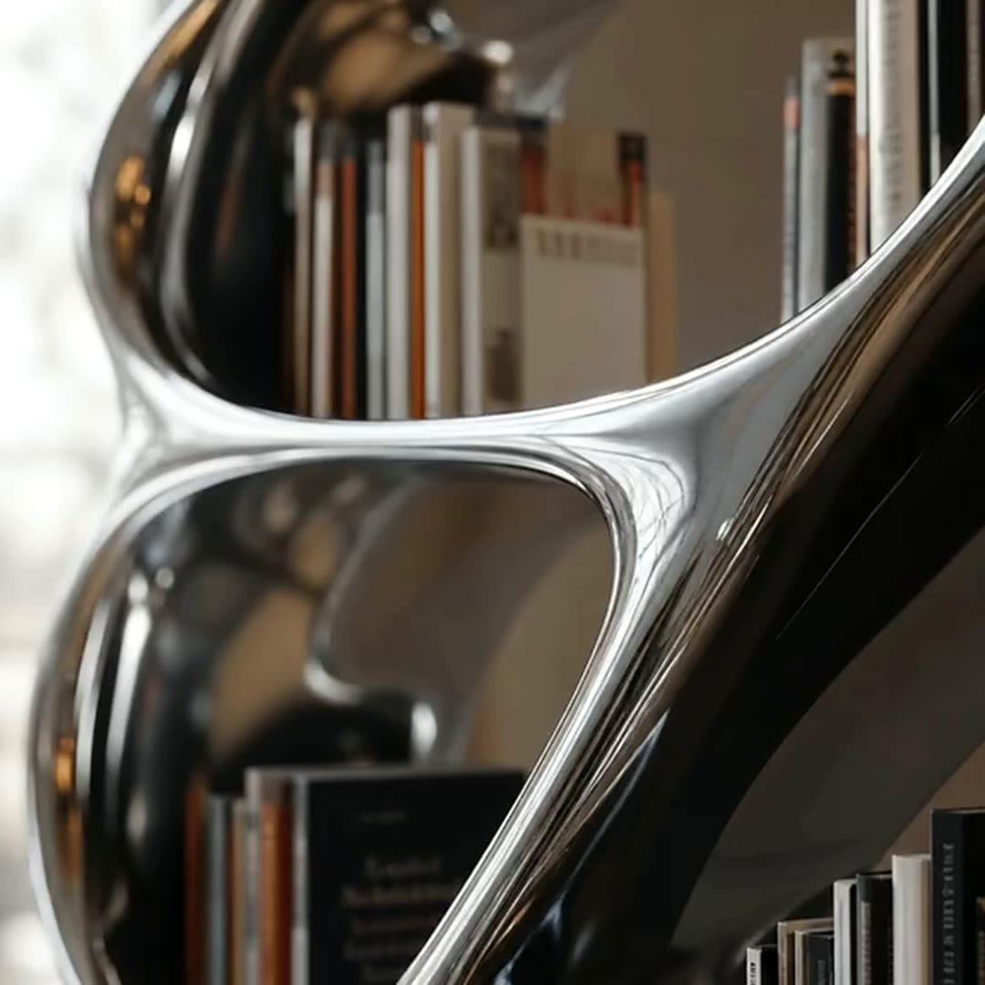 Close-up of books held inside the curved mirror-polished stainless steel openings, with the surrounding room reflected in the polished surface