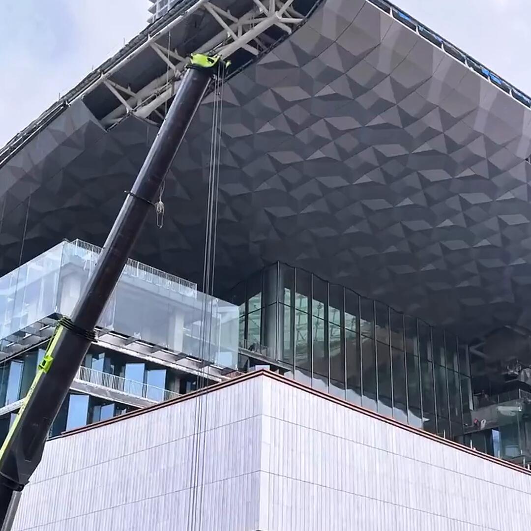 Daytime installation of polygonal stainless steel ceiling panels, a crane lifting a faceted panel into position against the structural frame of a shopping mall canopy