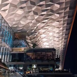 The finished polygonal stainless steel ceiling installed inside a shopping mall atrium at night, spanning four retail levels above escalators and crowds