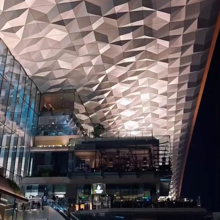 The finished polygonal stainless steel ceiling installed inside a shopping mall atrium at night, spanning four retail levels above escalators and crowds