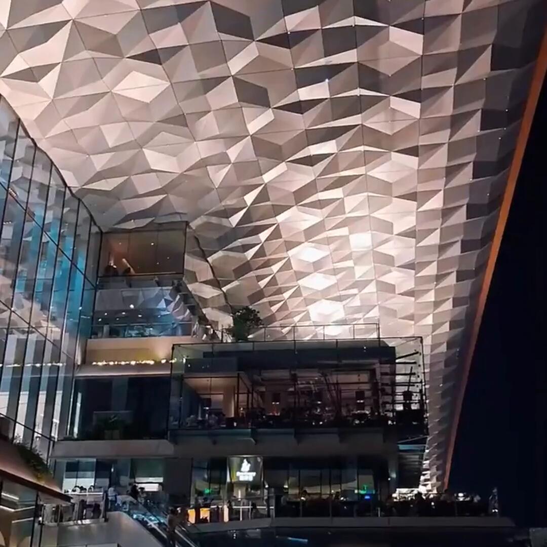The completed polygonal stainless steel ceiling installed in a shopping mall atrium at night, fold after fold of brushed metal lit across four retail levels above escalators and footfall