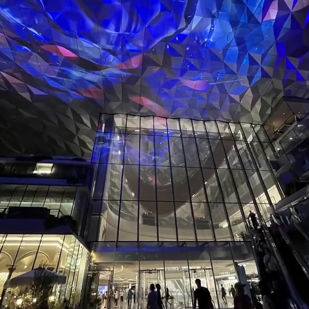 The same stainless steel ceiling running a mapped laser and projection show at night, with a swirling vortex image and blue wave imagery moving across the folded metal facets
