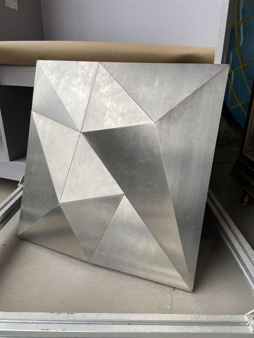 A completed polygonal stainless steel ceiling panel standing in the workshop, its brushed face folded into a low-poly field of triangular facets, with aluminium T-slot framing profiles lying in front of it