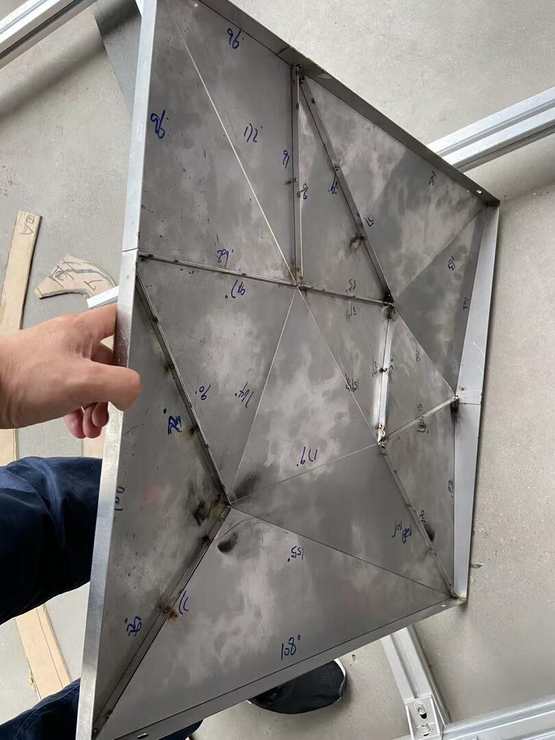 Reverse side of a polygonal stainless steel ceiling panel showing the welded stainless frame and triangular sub-structure, each facet numbered for installation, bolted onto aluminium T-slot framing