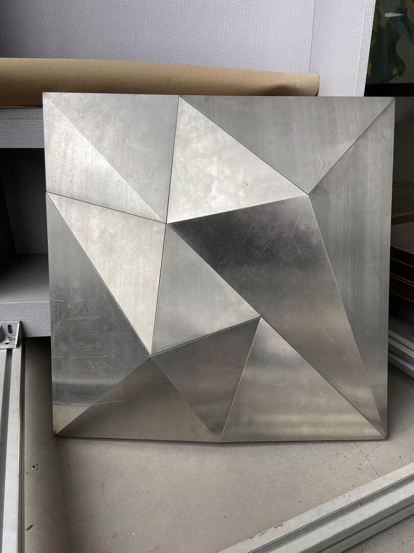 Close-up of the folded triangular facets of a brushed stainless steel ceiling panel, showing the crisp seam lines and the way each flat face catches light at a different angle