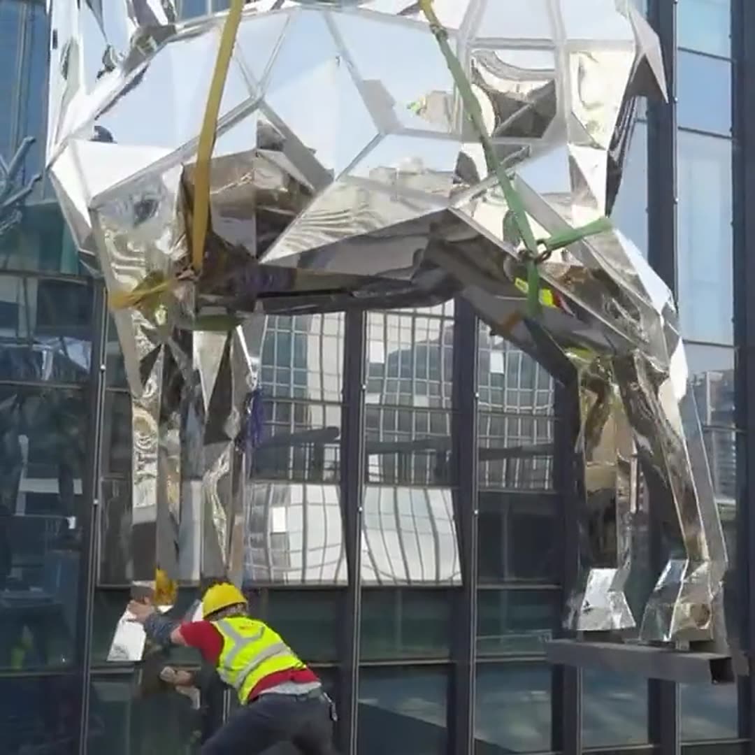 Faceted mirror-polished stainless steel deer lowered by crane slings against a glass curtain wall