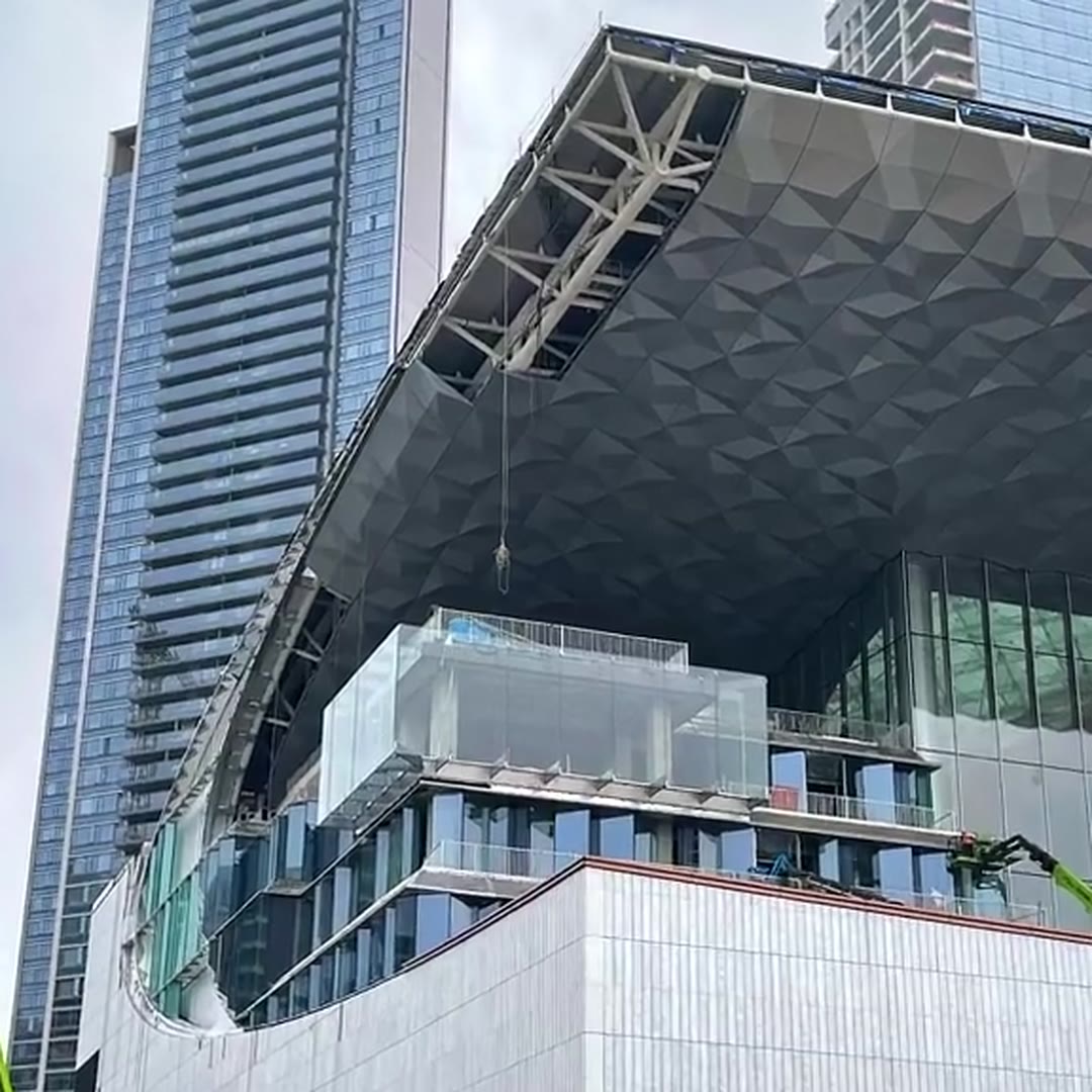 Cantilevered exterior canopy clad in 3D faceted stainless steel panels above a glass commercial building during construction
