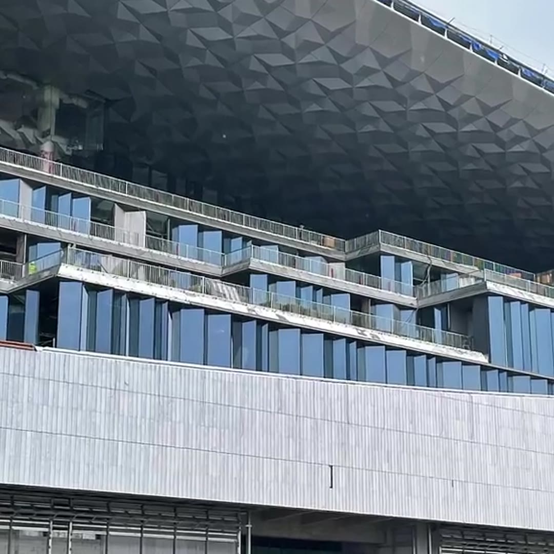Underside of a large faceted stainless steel canopy in daylight, showing the folded triangular facet pattern above a glass curtain wall