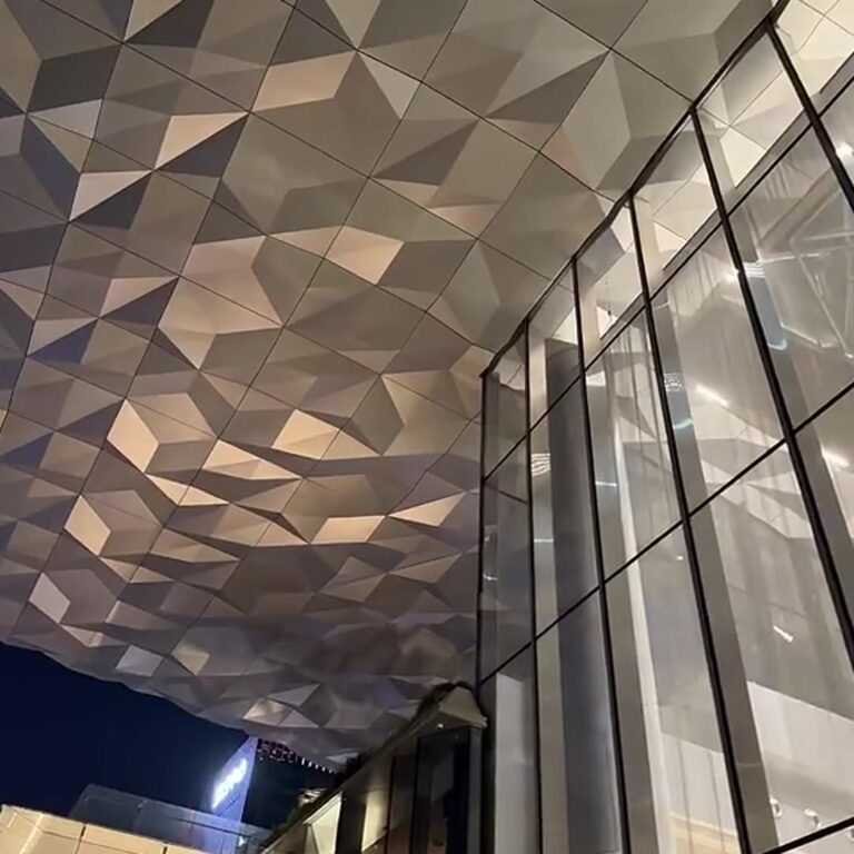 Indoor 3D faceted stainless steel ceiling lit by pin-spot fixtures at every vertex beside a full-height glass wall