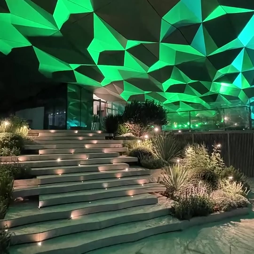 Outdoor stepped plaza with planted terraces and pin-spot garden lights under a faceted stainless steel canopy washed in green light at night