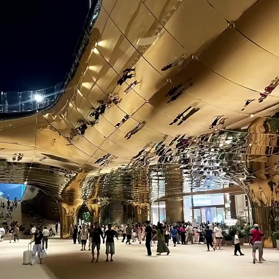 Night view of an undulating champagne-gold mirror stainless steel atrium canopy reflecting crowds of visitors and storefront lights below