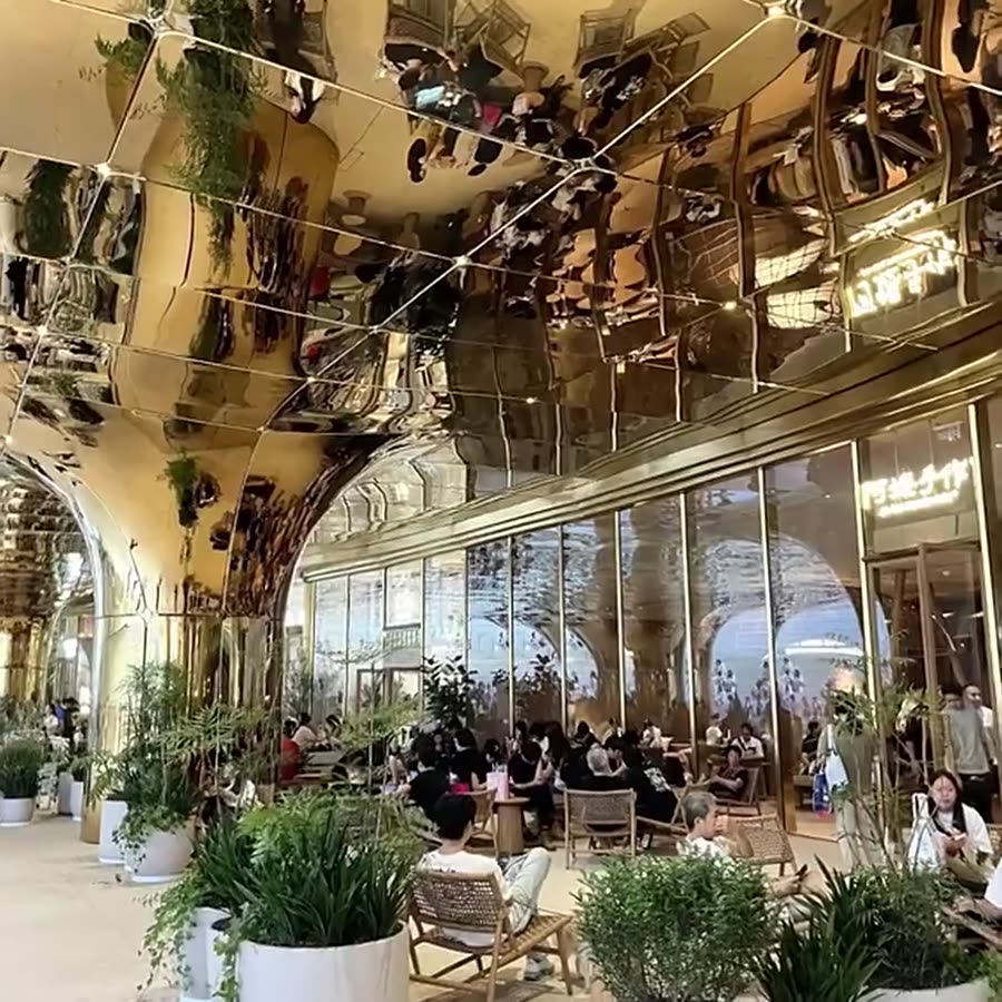 Champagne-gold mirror stainless steel wall and arched openings running along a mall corridor with ceiling-height cafe seating and glass shopfronts
