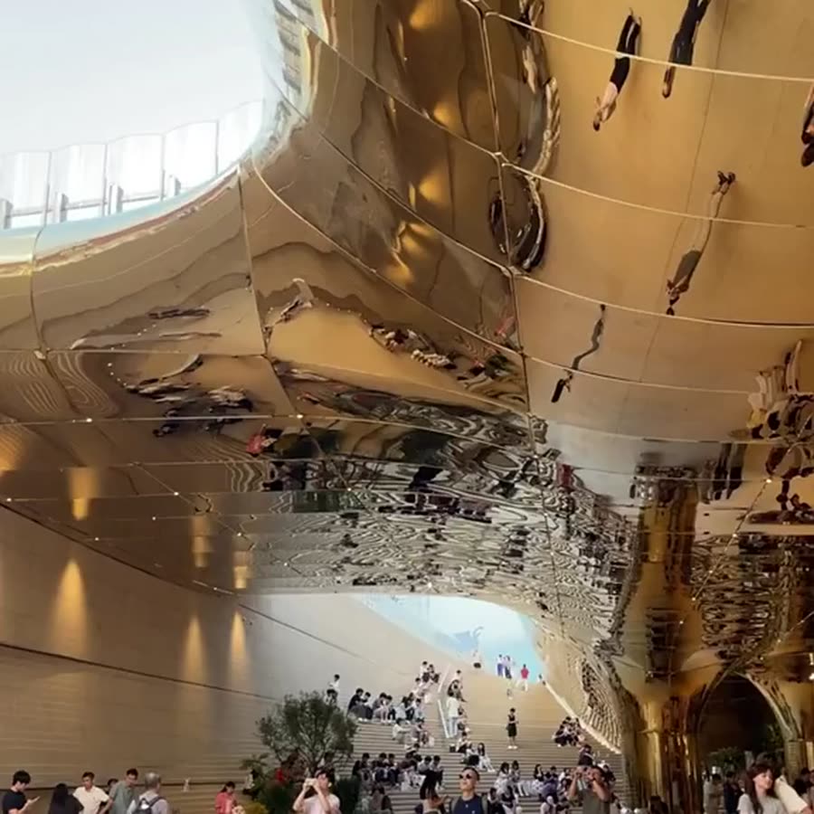 Sweeping curved champagne-gold mirror stainless steel canopy arching over a grand mall staircase beneath a skylight oculus