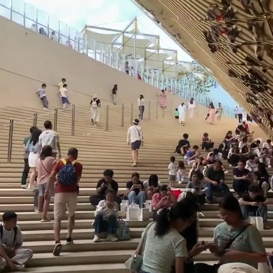 Wide atrium staircase with visitors resting on the steps, champagne-gold mirror stainless steel canopy edge visible overhead
