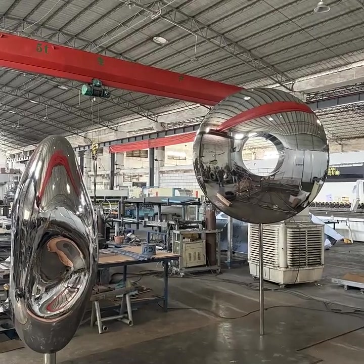 Two large mirror-polished stainless steel ring sculptures standing on steel support frames inside the ARTDSN fabrication workshop
