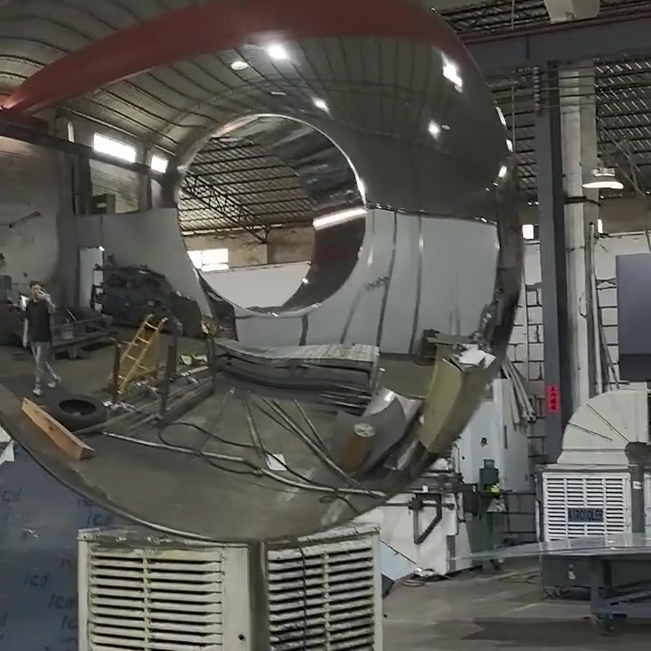 Closer view of the ring sculpture on its temporary steel jig, reflecting workers and equipment in the fabrication shop