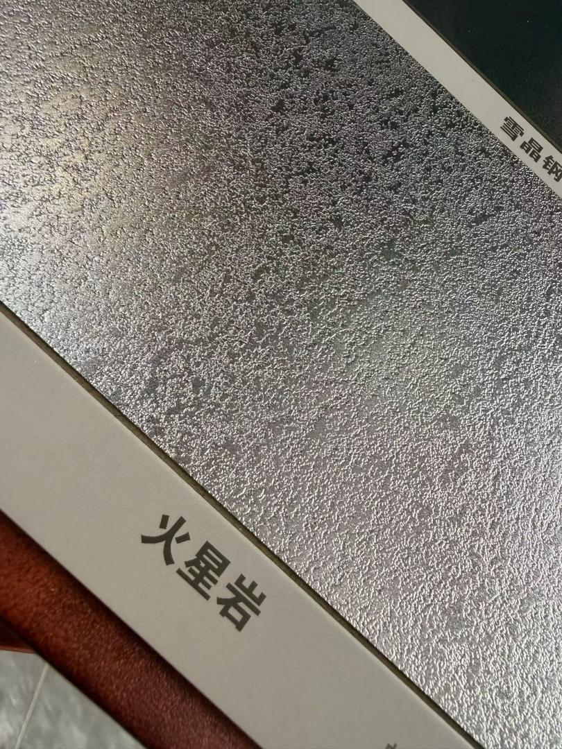 Close-up of a bright etched stainless steel sheet sample with a shimmering irregular anti-scratch pattern and a copper panel behind it