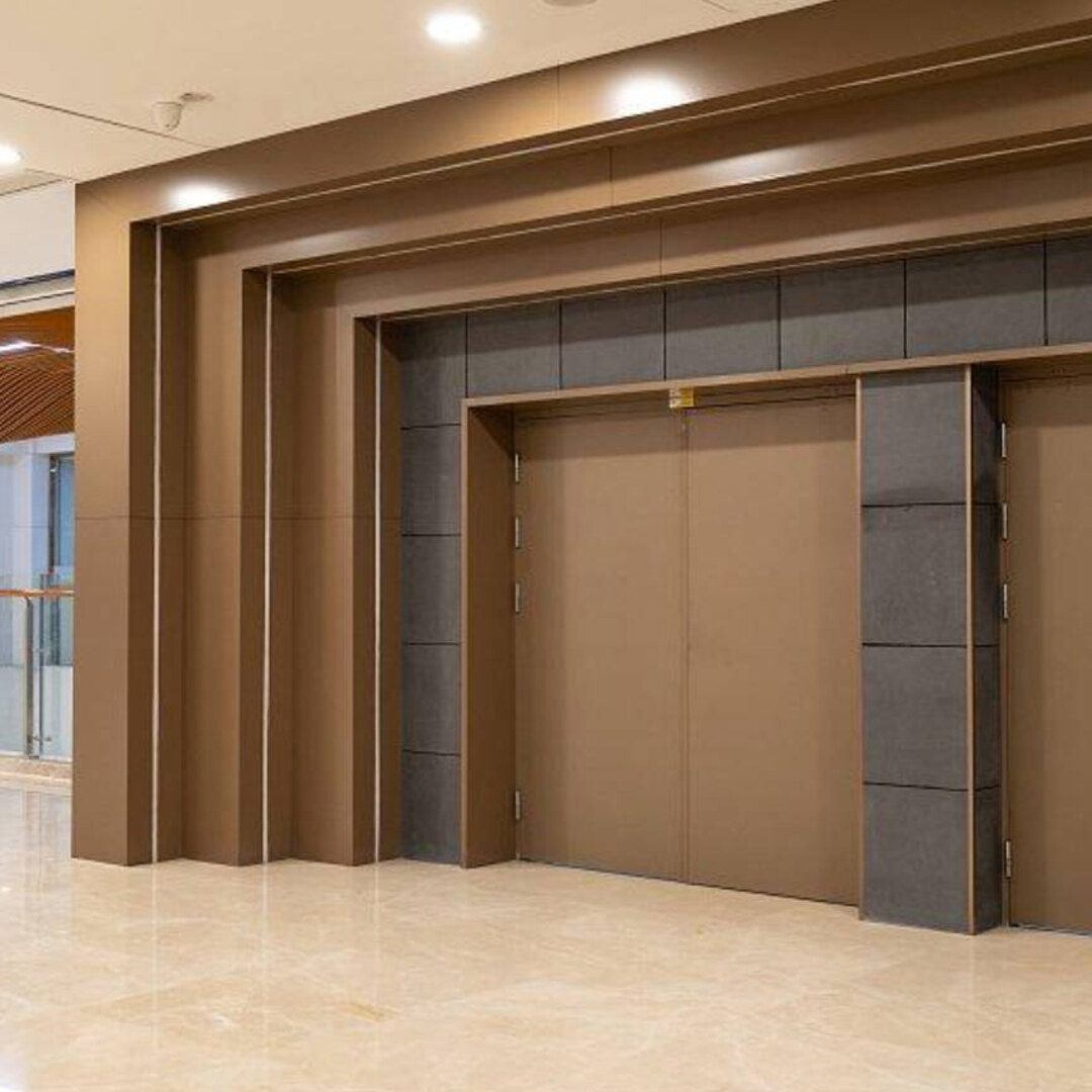 A hotel lobby interior where stainless steel cladding forms stepped portal frames around bronze-finished double entrance doors, with dark stone-look panels between the frames